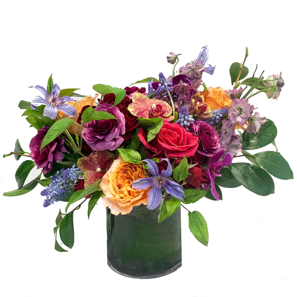 A whimsical vibrant garden-style arrangement using bright colored premium blooms accented with seasonal greenery in a chic glass vase.