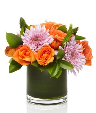 A mixed flower arrangement of lavender mums and orange roses arranged with greens in a glass vase.