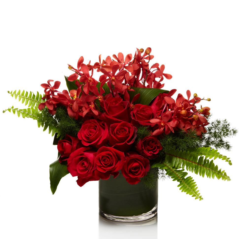 A lush arrangement of red roses and red orchid varieties, accented with modern greens in a premium glass vase.